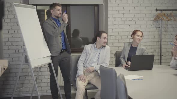 Bearded Caucasian Man in Grey Suit Standing Next To Board in Open Space Office and Talking with