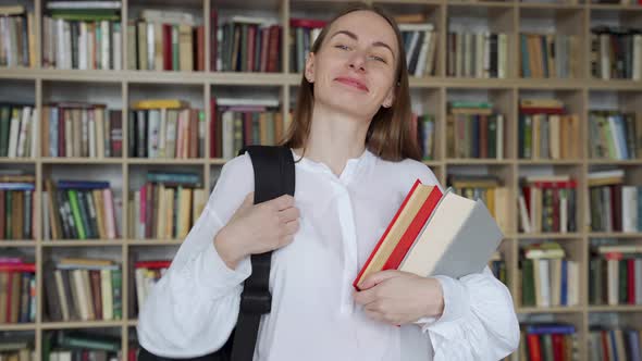 Young Smiling Woman Stands with Books and a Backpack in the Library