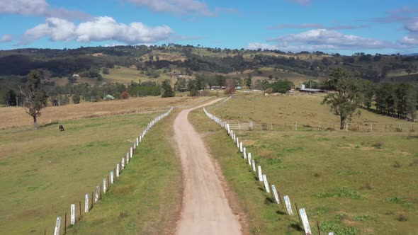 Aerial Footage of A Dirt Road with White Fence Posts Running Through a Green Farming Field