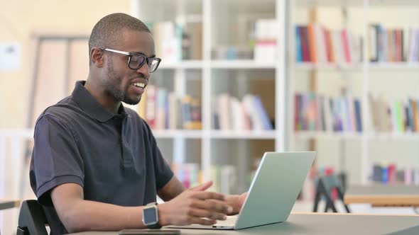 Cheerful Young African Man Doing Video Chat on Laptop in Library