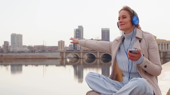 A Young Woman in Headphones and Listens to Relaxing Music on the Embankment