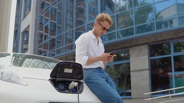 Stylish Modern Young Curly Man Standing Near an Electric Car Charging and Swiping in His Smartphone