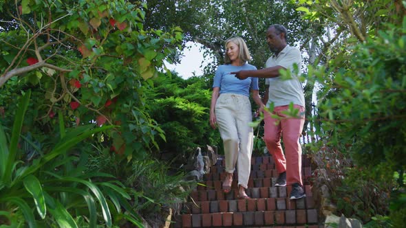 Diverse senior couple walking in their garden in the sun holding hands and talking