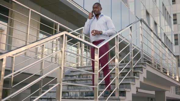 Afroamerican Businessman with Coffee Cup Talking on Smartphone Standing on Stairs Outside Office