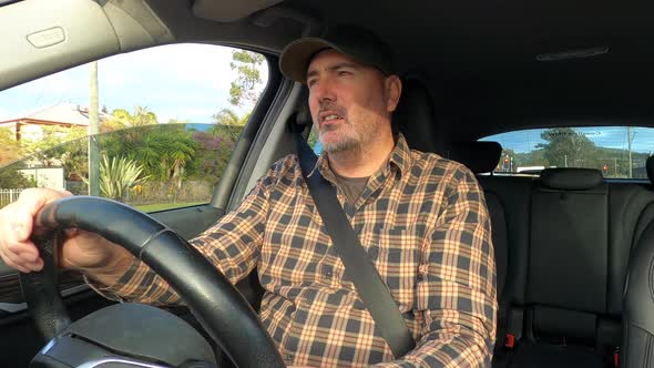 A man wearing a cap squints and uses a sun visor while driving in a country town towards the sun.