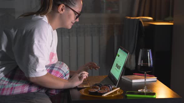 Woman in Pajamas and Glasses is Sitting on Sofa and Working on Laptop at Home