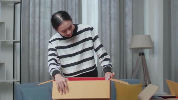 Young Asian Woman Moving Into A New House Unpacking The Cardboard Box And Taking Stuff Out Of It
