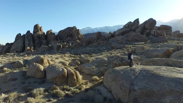 Aerial shot of a young man backpacker standing on a boulder with his dog in a desert mountain range.