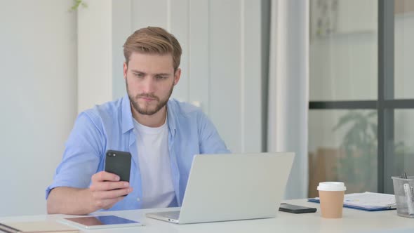 Young Creative Man with Laptop Using Smartphone at Work