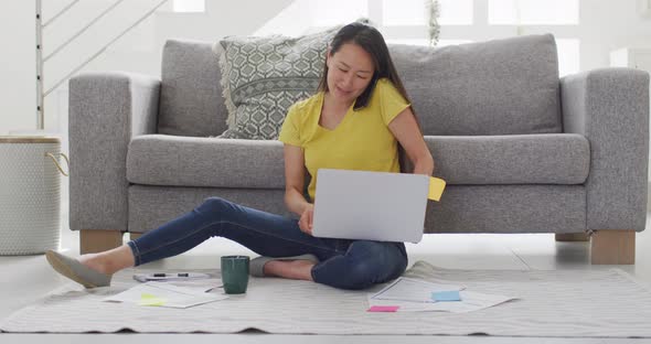 Focused asian woman sitting on floor and working remotely from home with laptop