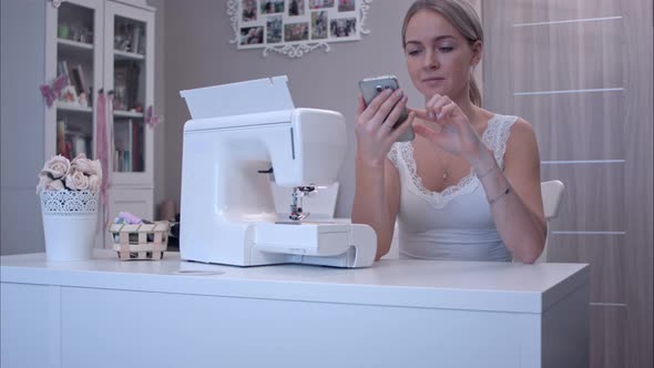 Young Woman with Phone Sitting at the Table Next To Sewing Machine