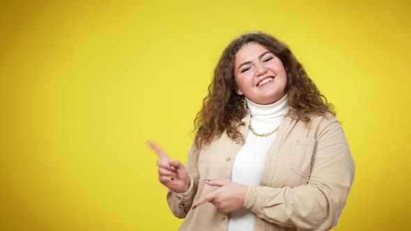 Joyful Smiling Plussize Woman Looking at Camera Pointing Aside Standing at Yellow Background