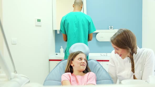 Smiling dentist talking to young female patient, male surgeon in background