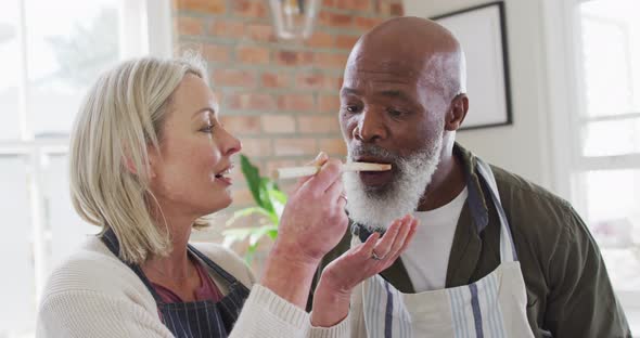 Mixed race senior couple wearing aprons tasting food while cooking in the kitchen at home