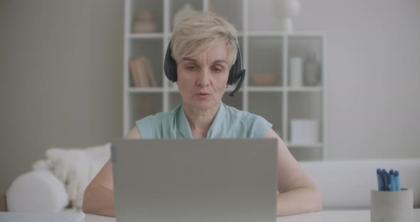 Aged Woman Is Communicating Online, Using Laptop with Internet and Headphones with Microphone