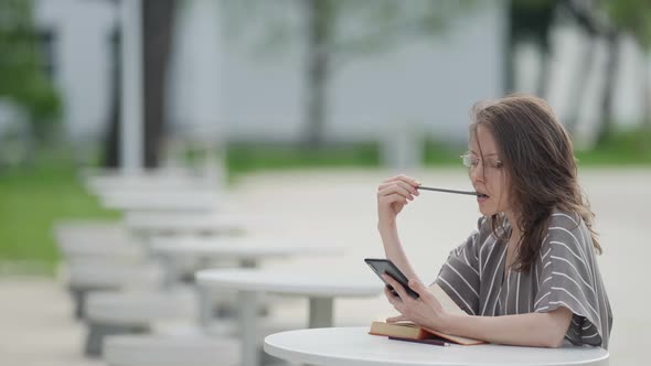 Clever Woman with Glasses is Reading Book and Surfing Internet By Smartphone Outdoors