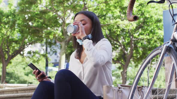 African american woman wearing face mask using smartphone drinking coffee in city park