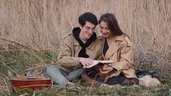 Young Couple Sitting in Field and Reading Book Aloud