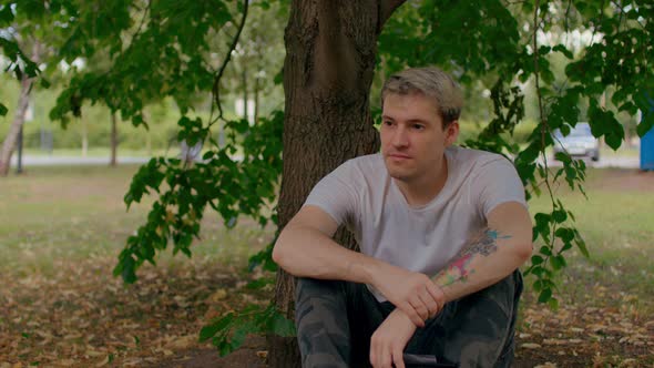 Close Up of Young Man Rests Sitting Near Tree in Parkland