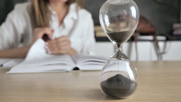 Pouring Sand in an Hourglass Standing on the Desk in Workspace. Woman Reading Books at Blur