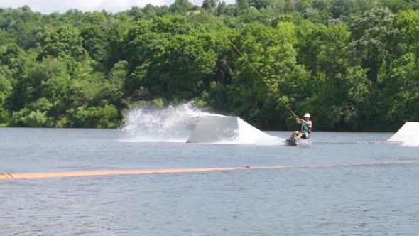 Athletic Man Wakeboarding and Doing Tricks Near Ramps on the Lake with Forest on Background