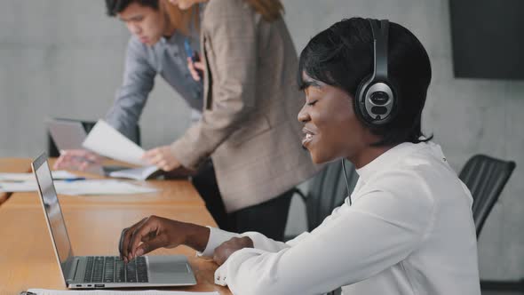 Smiling African Female Call Center Agent Wear Wireless Headset Using Laptop Looking at Screen