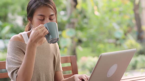 Freelance Asian woman working on laptop and drinking coffee sitting on table in the garden.