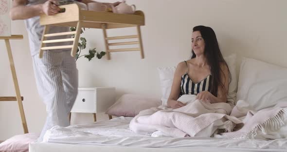 Brunette Caucasian Woman Lying As Handsome Husband Bringing Bed Tray with Tea, Croissants