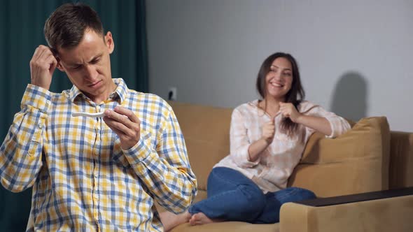 Young Man in Bewilderment Looks at a Pregnancy Test on the Background of a Happy Pregnant Woman Slow