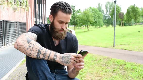 Young bearded tattooed man outdoor in the city using smart phone hand hold