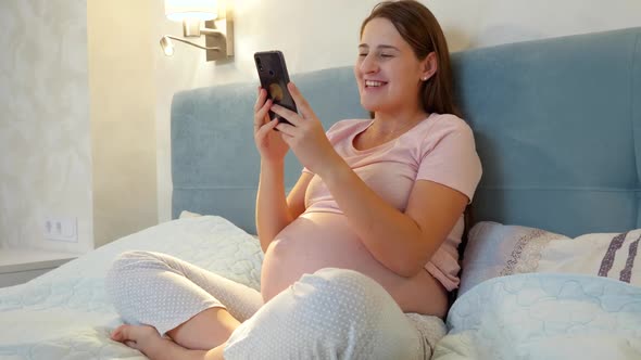 Young Pregnant Woman Talking on Video Call While Sitting on Bed at Night