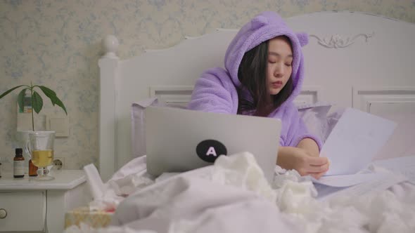 Sick 20s Asian Woman Working with Documents and Laptop Lying in Bed