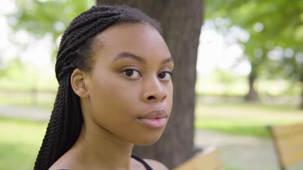 A Young Black Woman Turns and Looks Seriously at the Camera As She Sits on a Bench in a Park