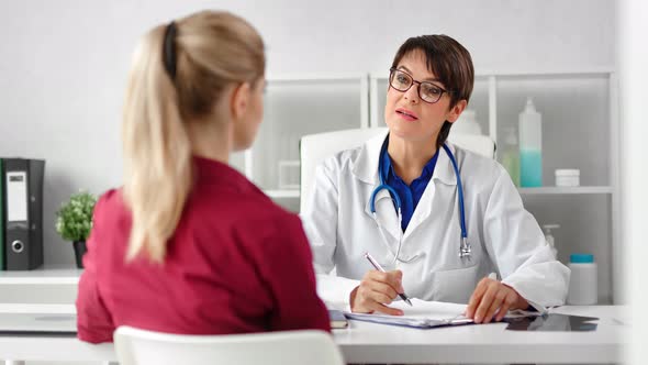 Woman Medical Worker in Glasses and Coat Talking with Girl Patient Taking Note During Consultation