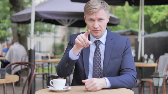 Outdoor Sitting Businessman Pointing with Finger Toward Camera