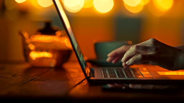 Female Fingers Typing on Laptop in Semidark Warm Setting. Bright Yellow Round Bokeh Background