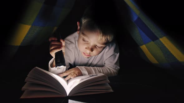 Boy reading a book with a flashlight under the covers at night alt