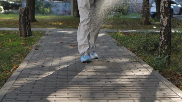 Bottom View of a Virologist in a White Protective Suit Disinfects Paving Slabs in a Public Park. The