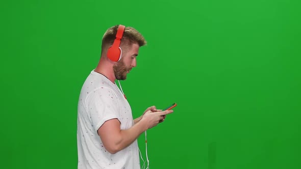Portrait Bearded Guy in Red Headphones Walking and Listening Music Vie Phone at Greenscreen. Profile