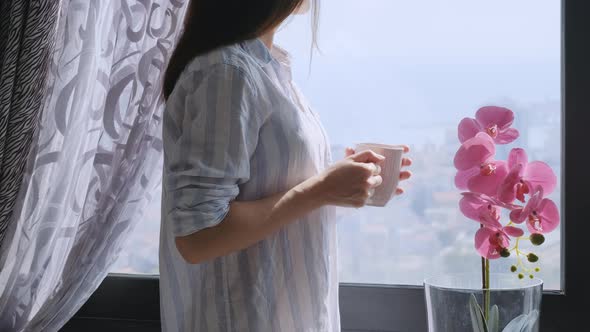 Millenial brunette woman holding a cup of tea or coffee and looking in the window