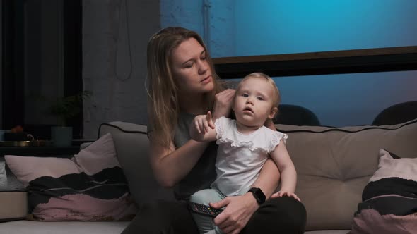 Cute Baby Girl and Young Mother Are Sitting on Gray Sofa with Black Remote Control in Hands and