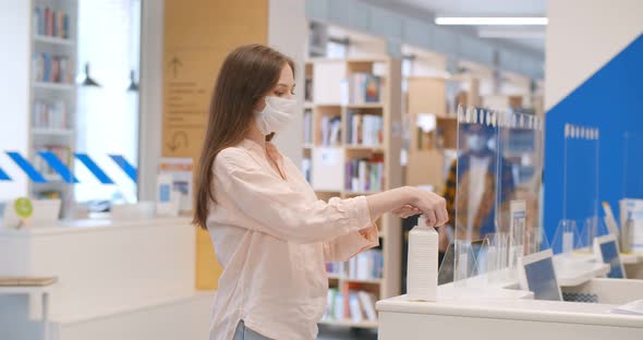 Female Student Wearing Protective Mask Disinfecting Hands with Antiseptic Gel on Desk in Library