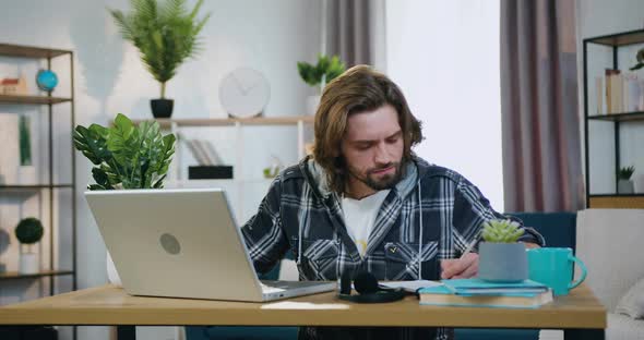 Man Sitting at the Desk at Home and Working with Computer and Papers