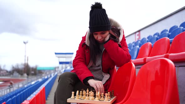 Young Woman in Winter Clothes Plays Chess Sitting on Stadium Bleachers Alone