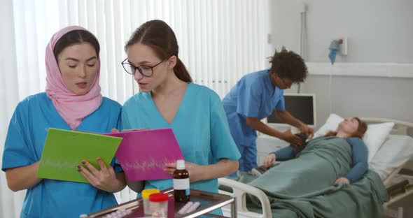 Two Women Doctors Looking at Clipboard with Colleague Treating Patient in Bed on Background