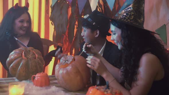 Two Girls and a Guy in Creepy Halloween Costumes are Sitting at a Table Chatting and Carving Faces