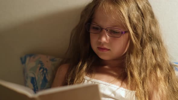 Child Girl in Glasses Reading Book in Bed Before Going to Sleep with Night Lamp