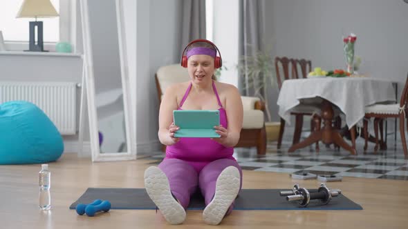 Excited Obese Woman Laughing Out Loud Sitting on Exercise Mat Watching Comedy Movie Online on Tablet