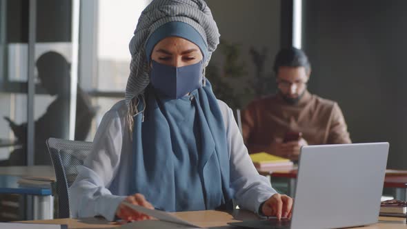Businesswoman in Hijab and Mask Using Laptop and Taking Notes