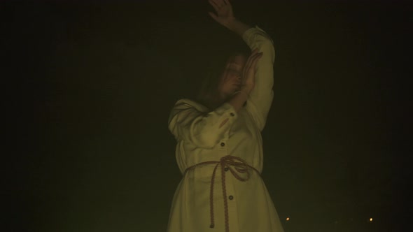 Slow ecstatic dance of young lady in white dress in darkness before fire yellow light low angle view